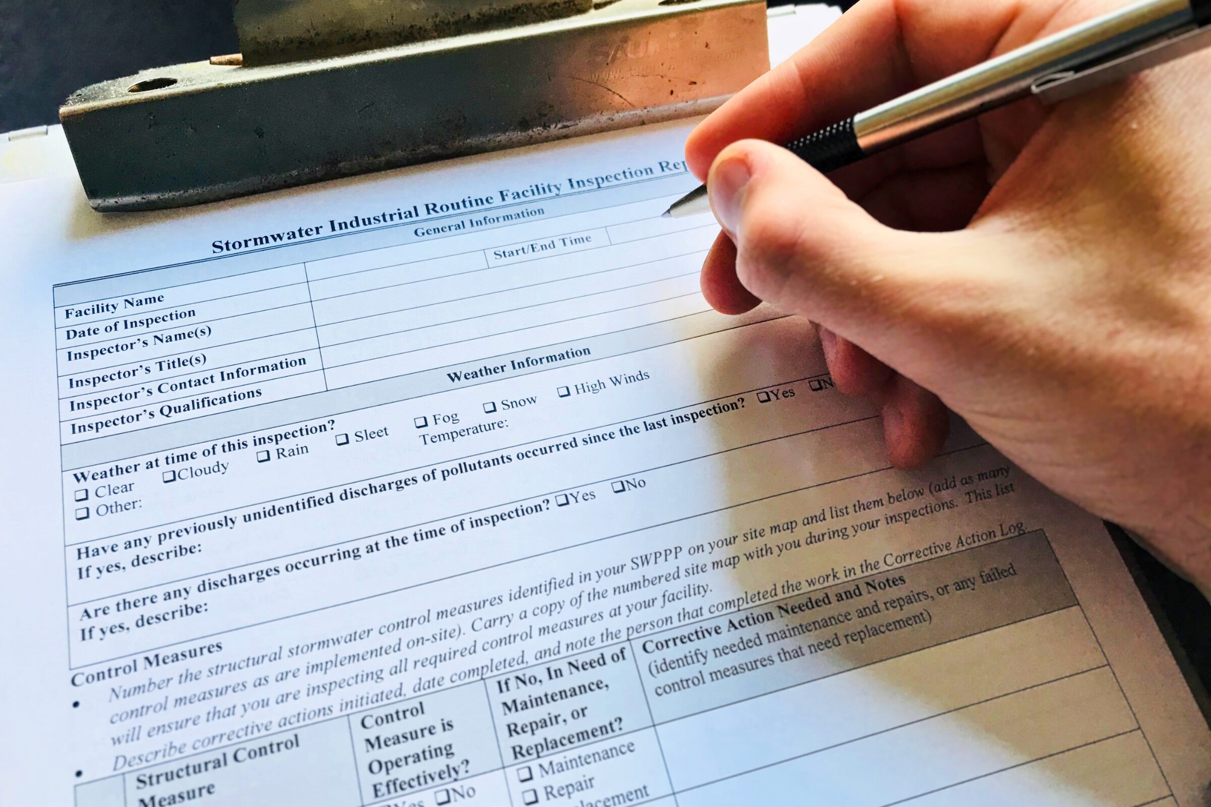 This image shows a close-up of a hand holding a pen and filling out a Stormwater Industrial Routine Facility Inspection Report on a clipboard. The form includes fields for facility name, date of inspection, inspector information, weather conditions, and notes on discharges or control measures. The inspection appears to be part of a SWPPP (Stormwater Pollution Prevention Plan) compliance process, typically required for facilities under NPDES permits. The scene captures an active, hands-on compliance check, suitable for use in environmental consulting or EHS-related communications.