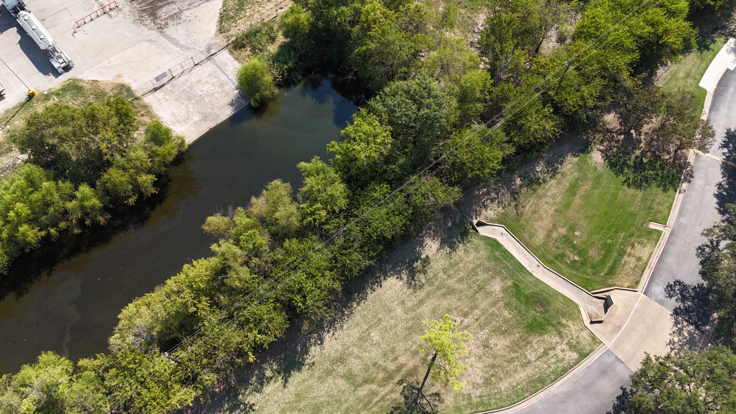An aerial photo showing a stormwater conveyance system in an industrial or commercial setting. A concrete outfall channel extends from a curbed parking lot or roadway, guiding stormwater runoff toward a naturalized stormwater retention pond. The receiving area is surrounded by dense vegetation and trees, which help stabilize the banks and provide erosion control. The pond is adjacent to a concrete pad and industrial facility with visible tanker trailers and paved surfaces.
