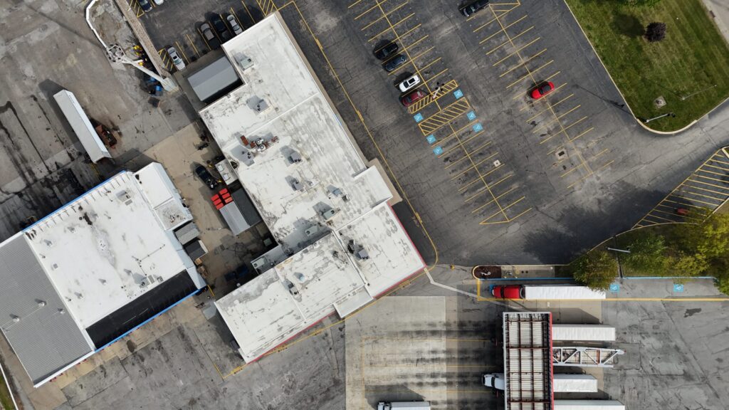 Drone image of an industrial facility with white membrane rooftops, truck loading areas, and parking lots, used to inspect stormwater drainage paths and evaluate potential spill risks for environmental compliance.